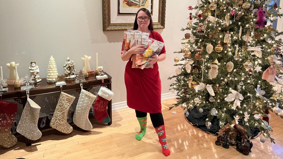 Jessica Rotolo holds socks bearing her winning design while wearing a pair of the socks and standing next to a Christmas tree.