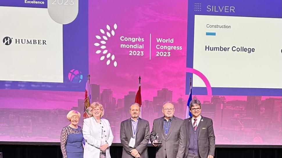 Five people, including Humber College president and CEO Ann Marie Vaughan, stand on a stage. One is holding an award.