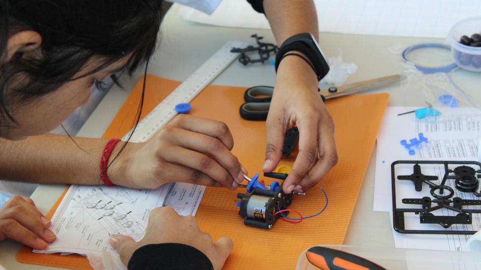 A person adjusts a small mechanical device with a tool with another person reads technical documents.