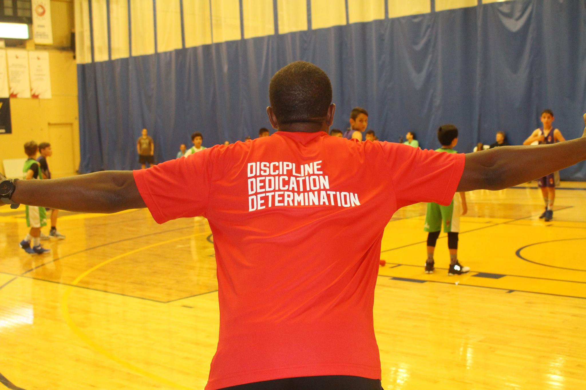 A person stands in a gym with their arms outstretched while youth play basketball in the background.