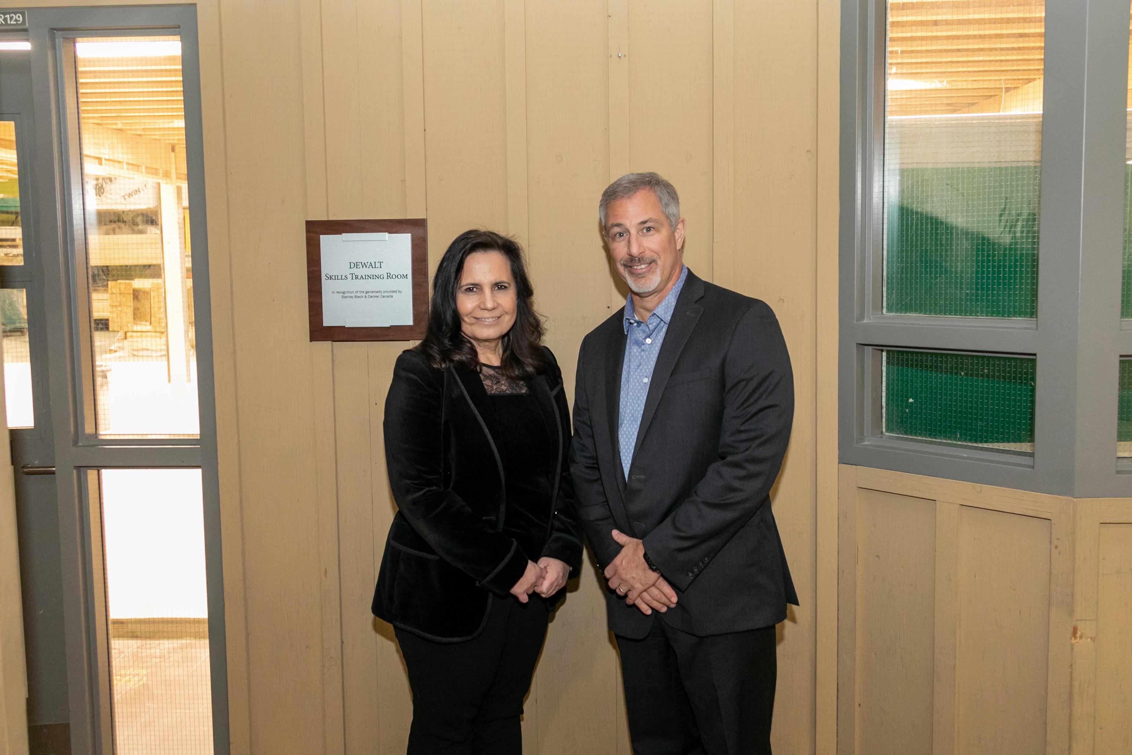 Two people smile and pose for a photo beside a sign that reads DEWLT Skills Training Room.