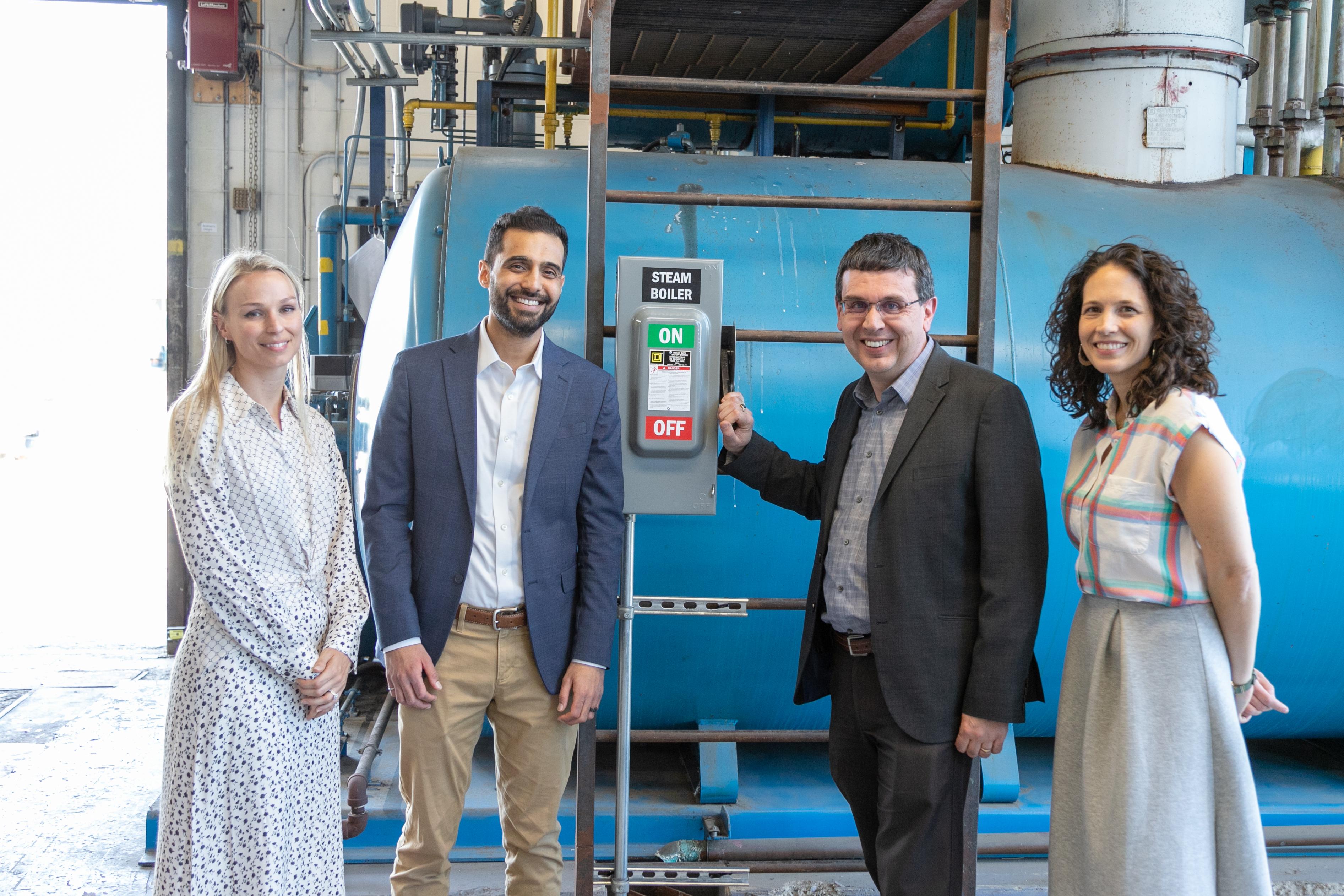 Four people stand in front of a large mechanical device. A lever that reads Steam Boiler is switched to off.