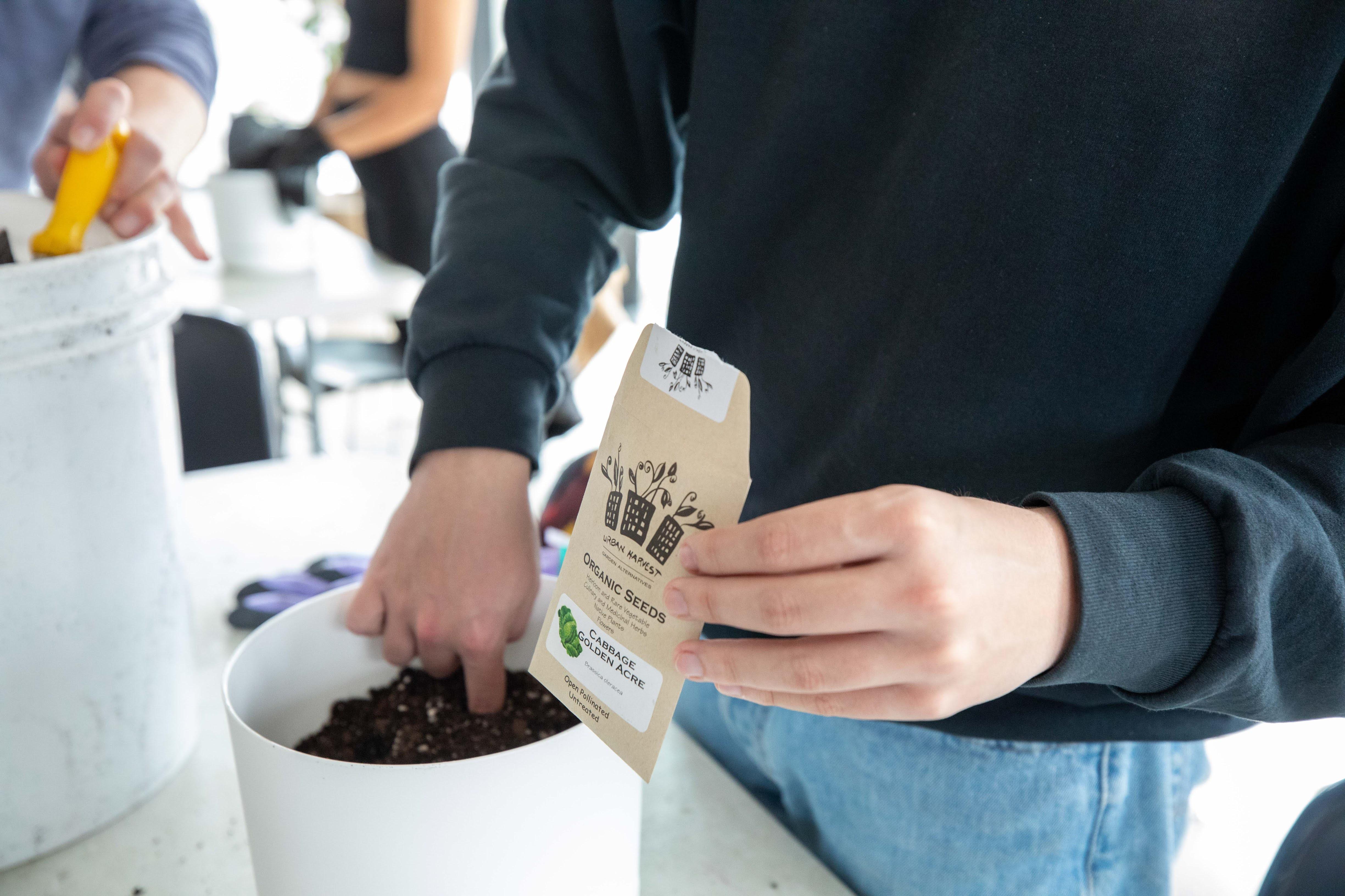 A person holds a packet of seeds while placing their finger in a pot of soil.