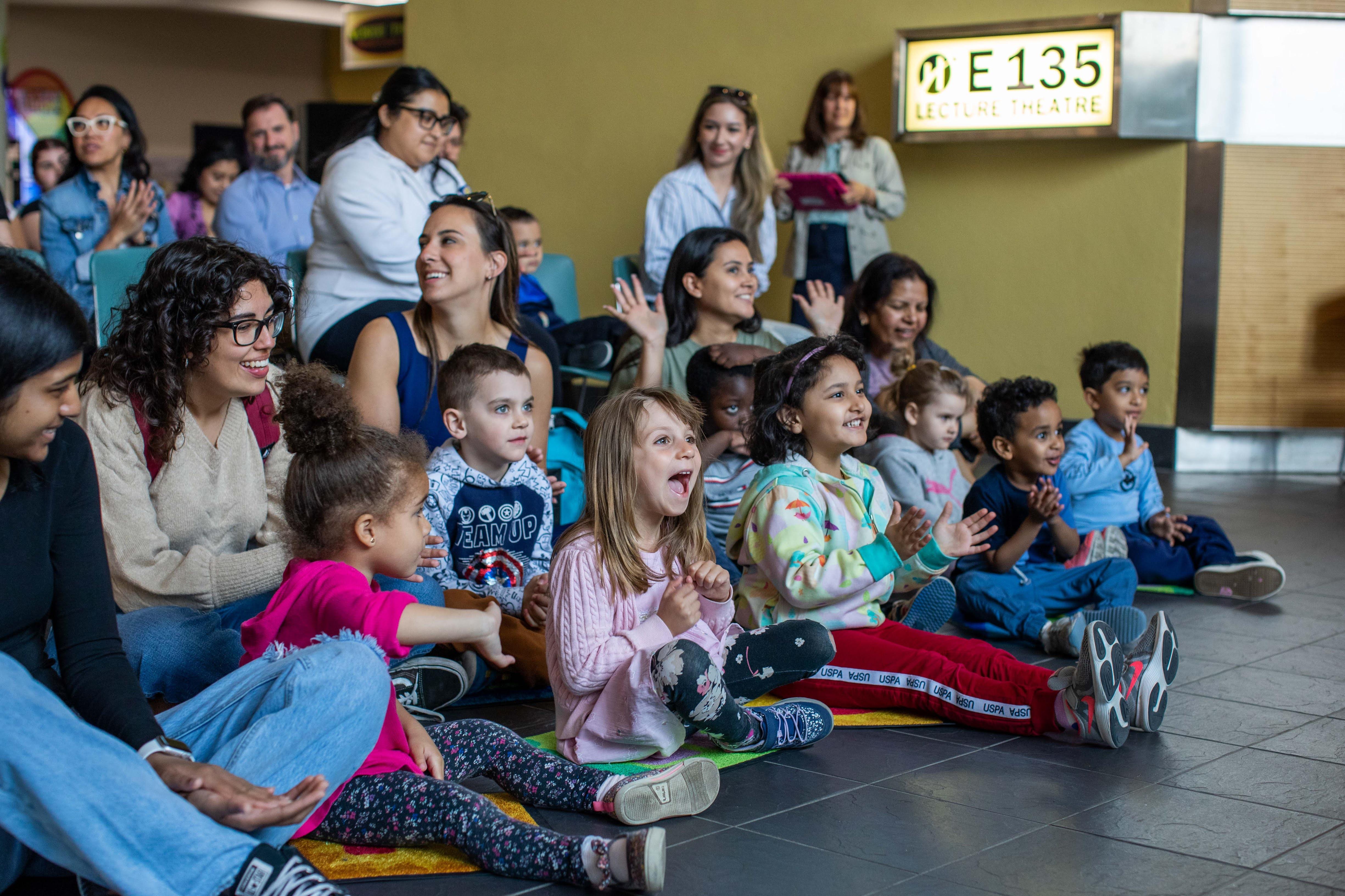 A group of children and parents sit together on the floor. They are smiling and clapping.