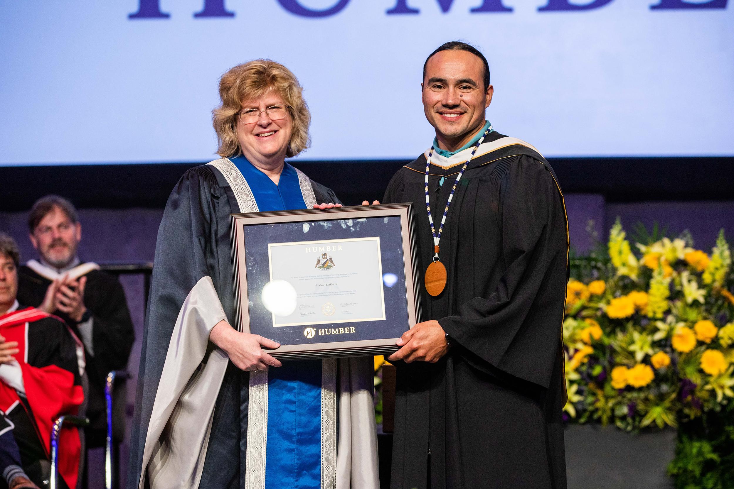 A person is presented with an honorary degree by another person while standing on stage.