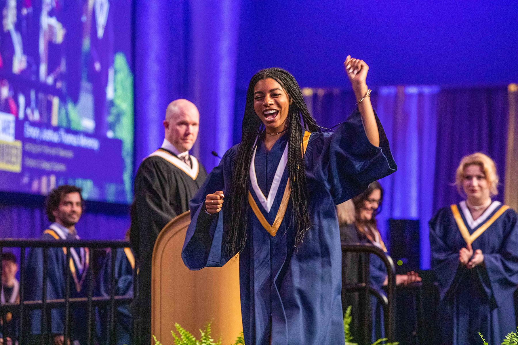 A person wearing graduation robes raises their arm and cheers as they walk across a stage.