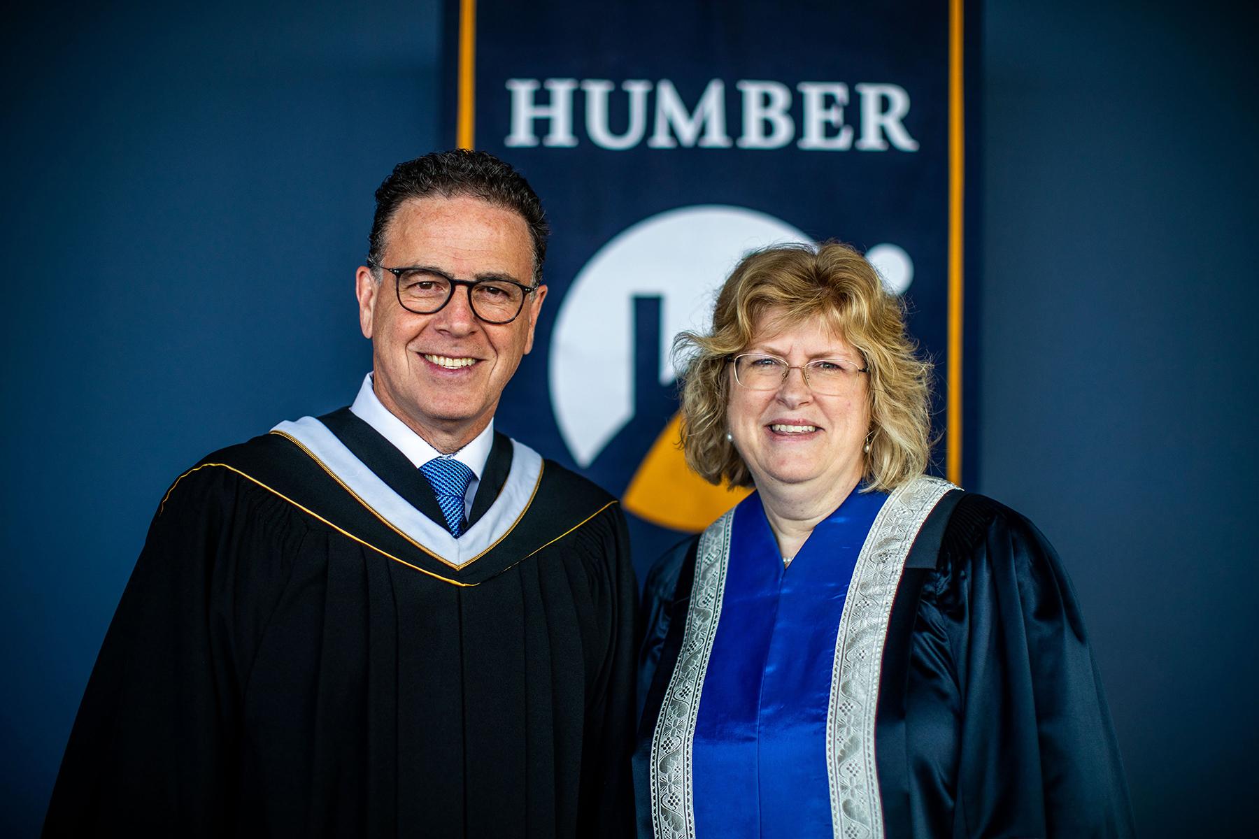 Two people wearing graduation robes smile and pose for a photo in front of a banner that reads Humber.