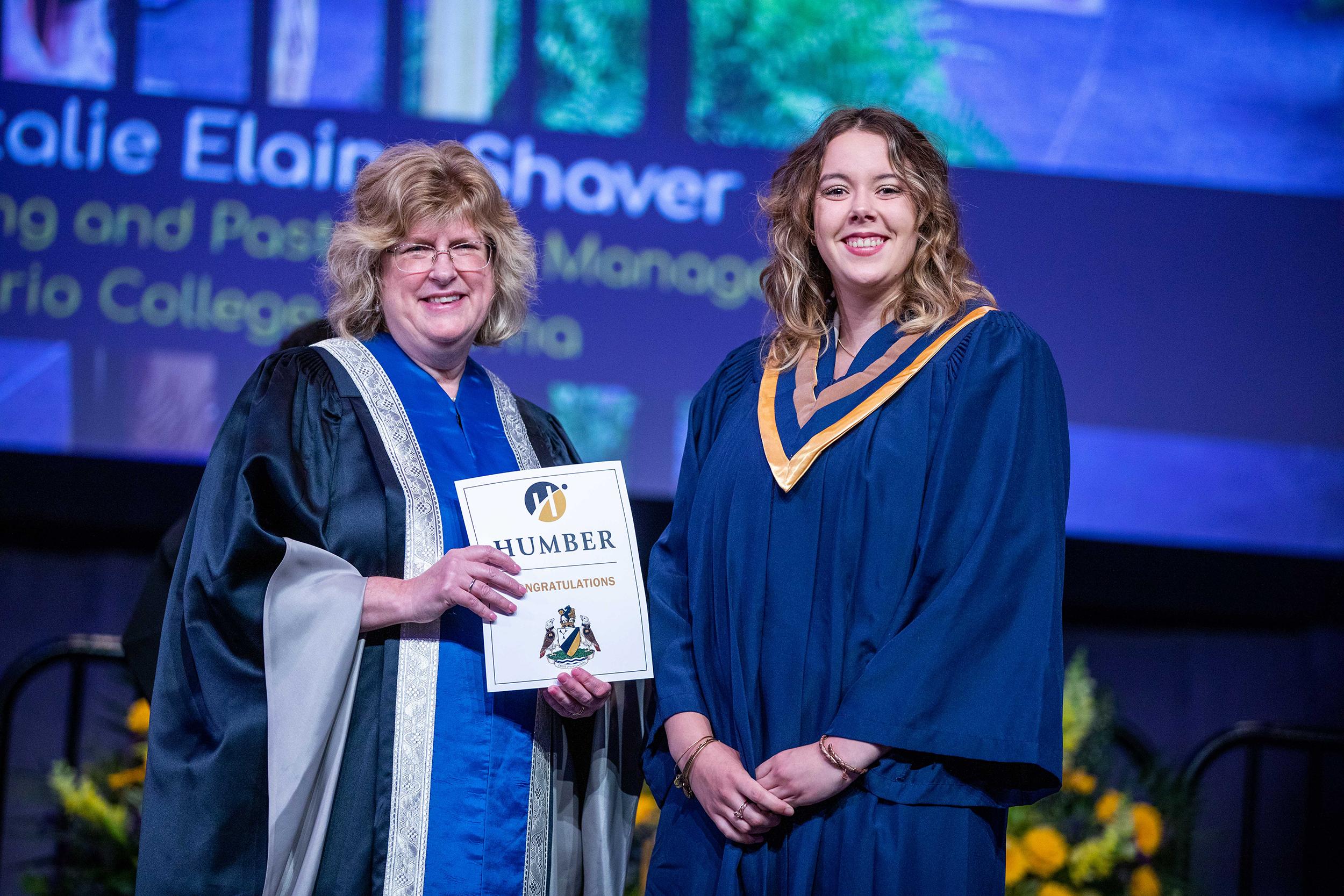 Two people wearing robes stand on a stage. One is holding a piece of paper that reads Humber Congratulations on it.