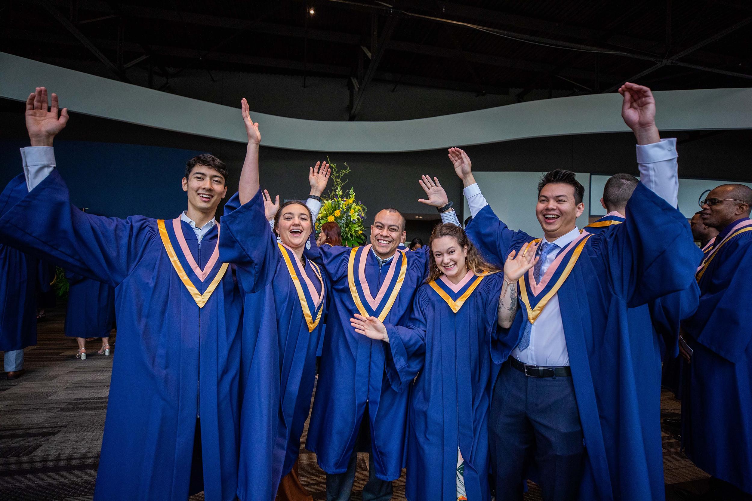 A group of people wearing graduation robes cheer and hold their arms above their heads in celebration.