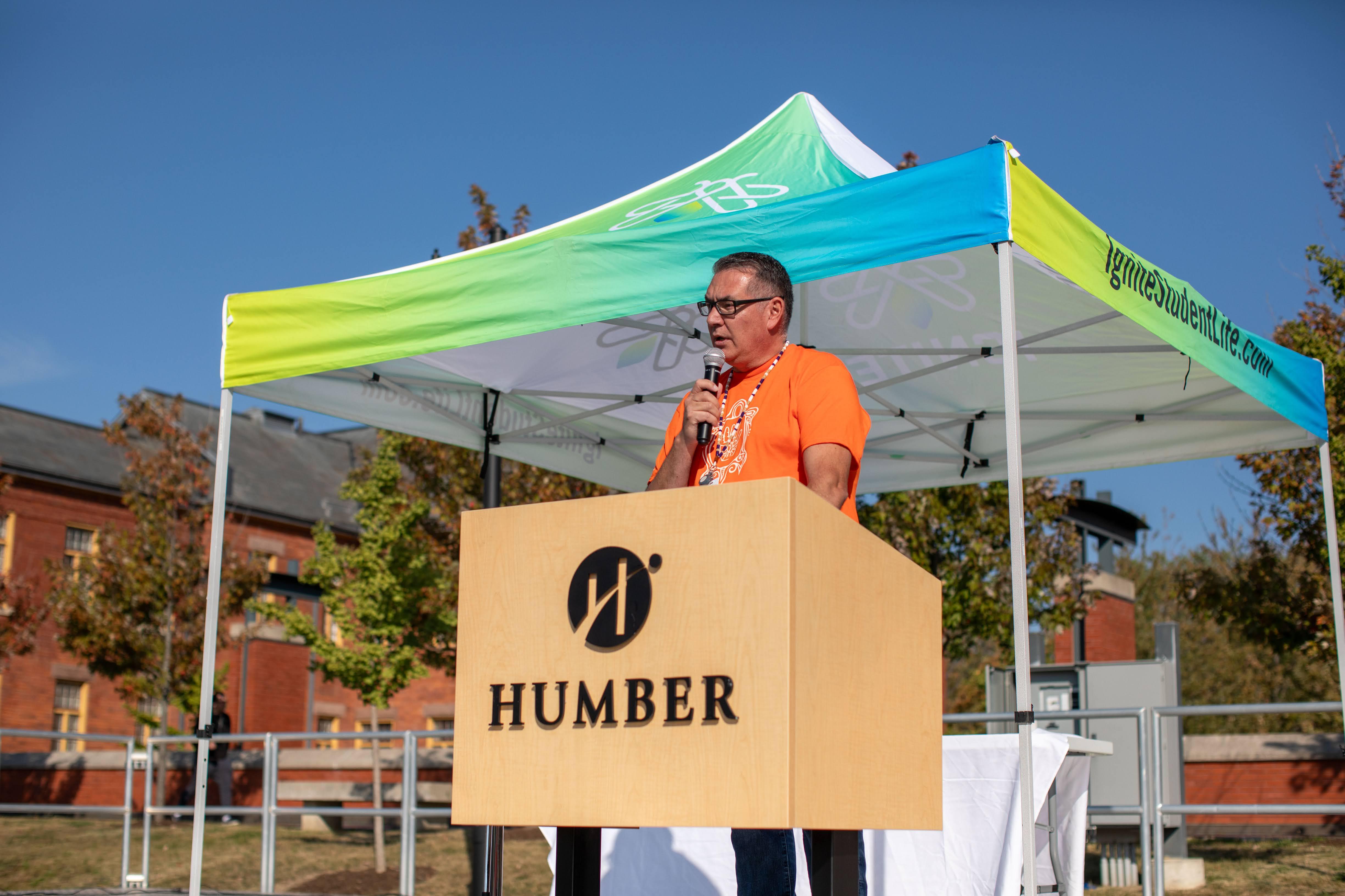 A person wearing an orange shirt speaks into a microphone while standing at a podium with Humber on it.