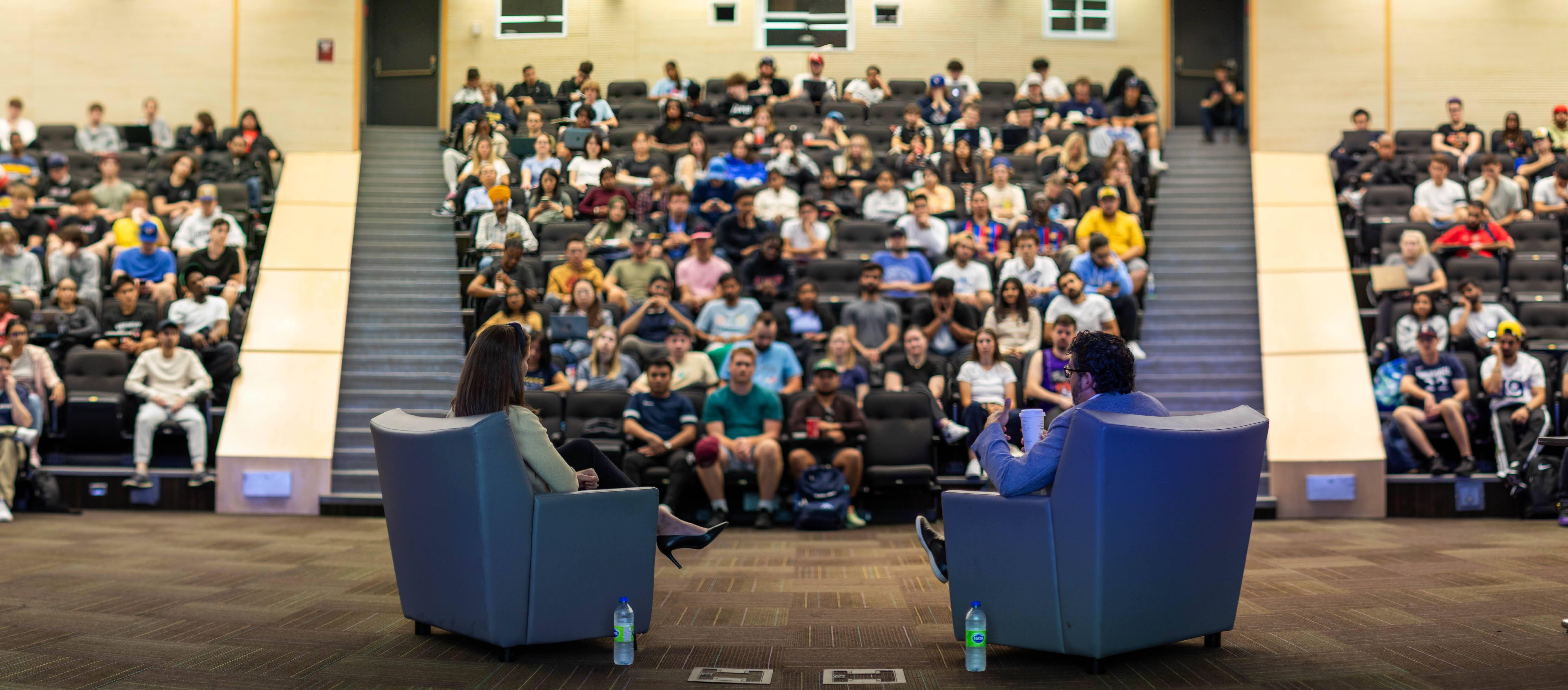 Two people sit in chairs while looking out towards the audience inside an auditorium.