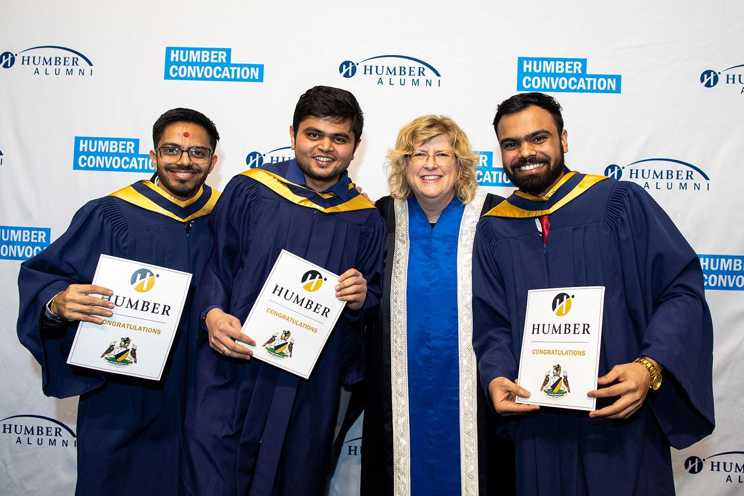 Three people wearing graduation gowns and holding pieces of paper pose for a photo with Humber College’s president and CEO.