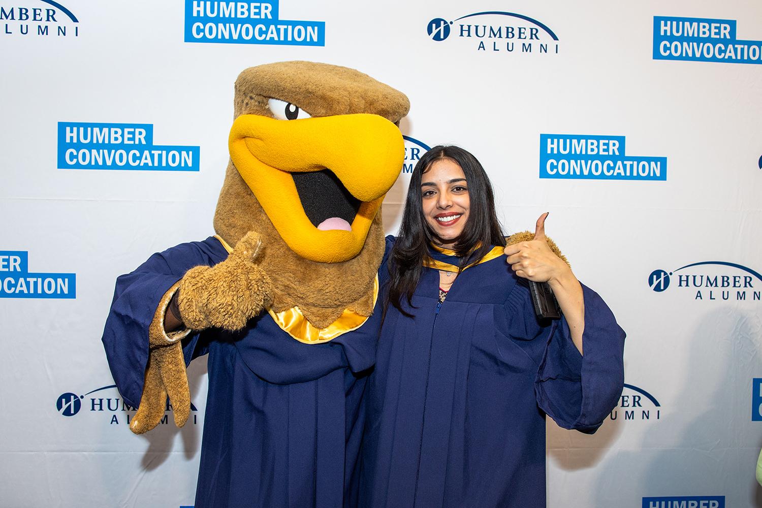 The Humber Hawk mascot poses for a photo giving a thumb’s up with another person wearing a graduation gown. 