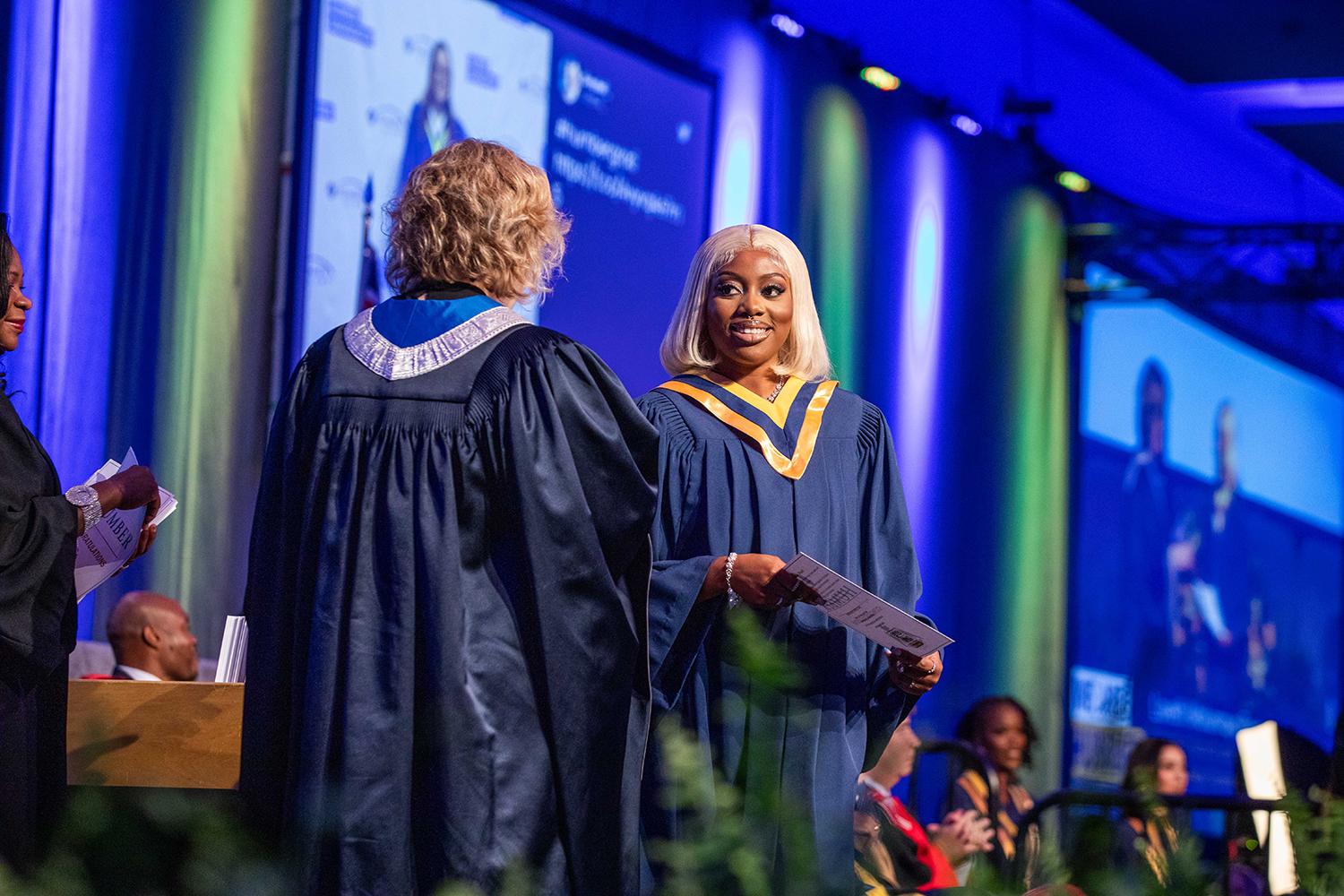 A person wearing a graduation gown holds a piece of paper in their hand as they walk across a stage.