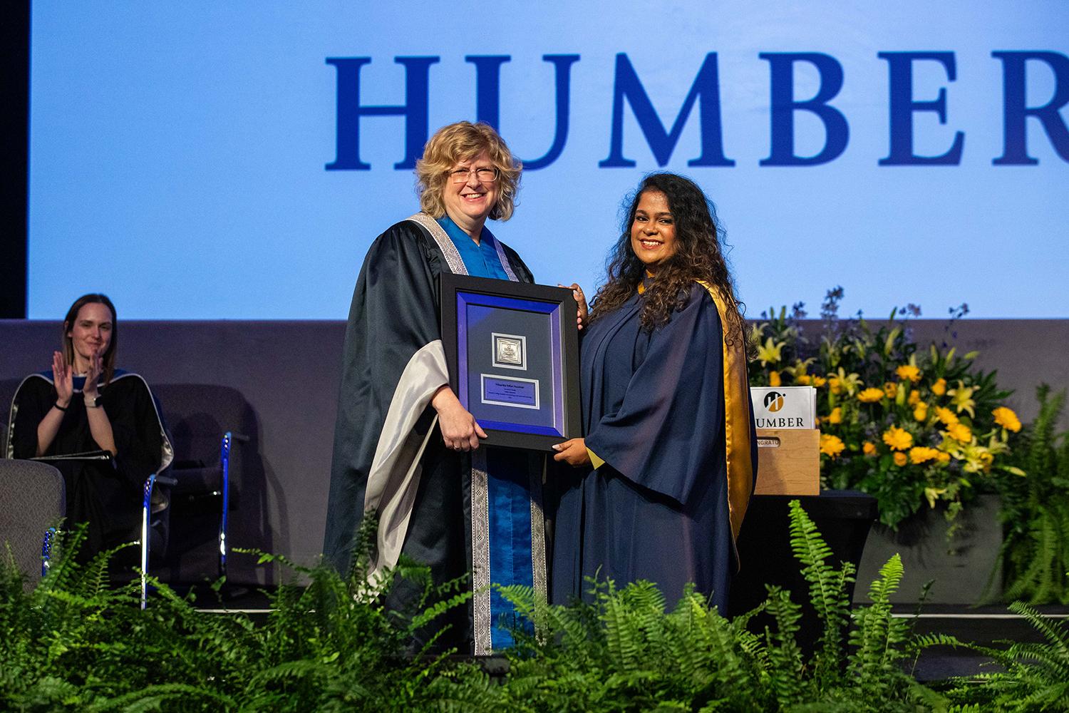 A person presents a student wearing a graduation gown with a framed credential. A screen reading Humber is in the background.