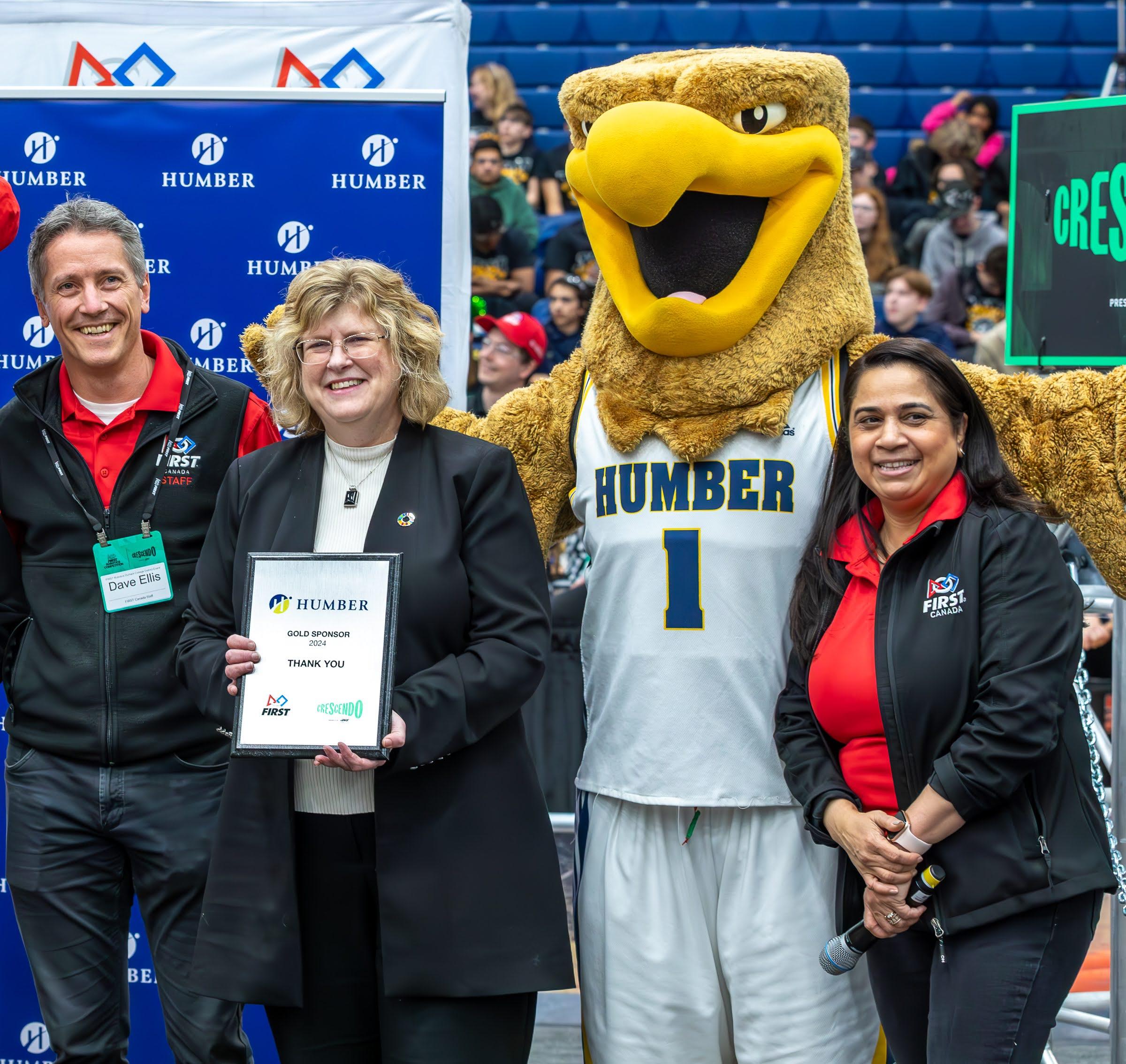 Three smiling people stand together along with the Humber Hawk mascot. One of them is holding up a plaque.