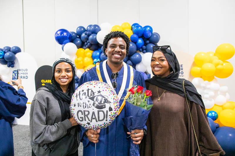 Three people stand together. One is wearing a graduation gown and is holding a balloon and flowers.