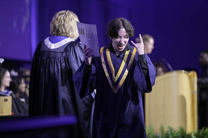 A person wearing a graduation gown and holding a credential celebrates on stage.