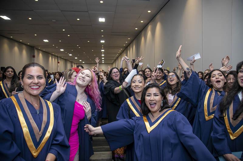 A large group of smiling people wearing graduation gowns cheer and wave.