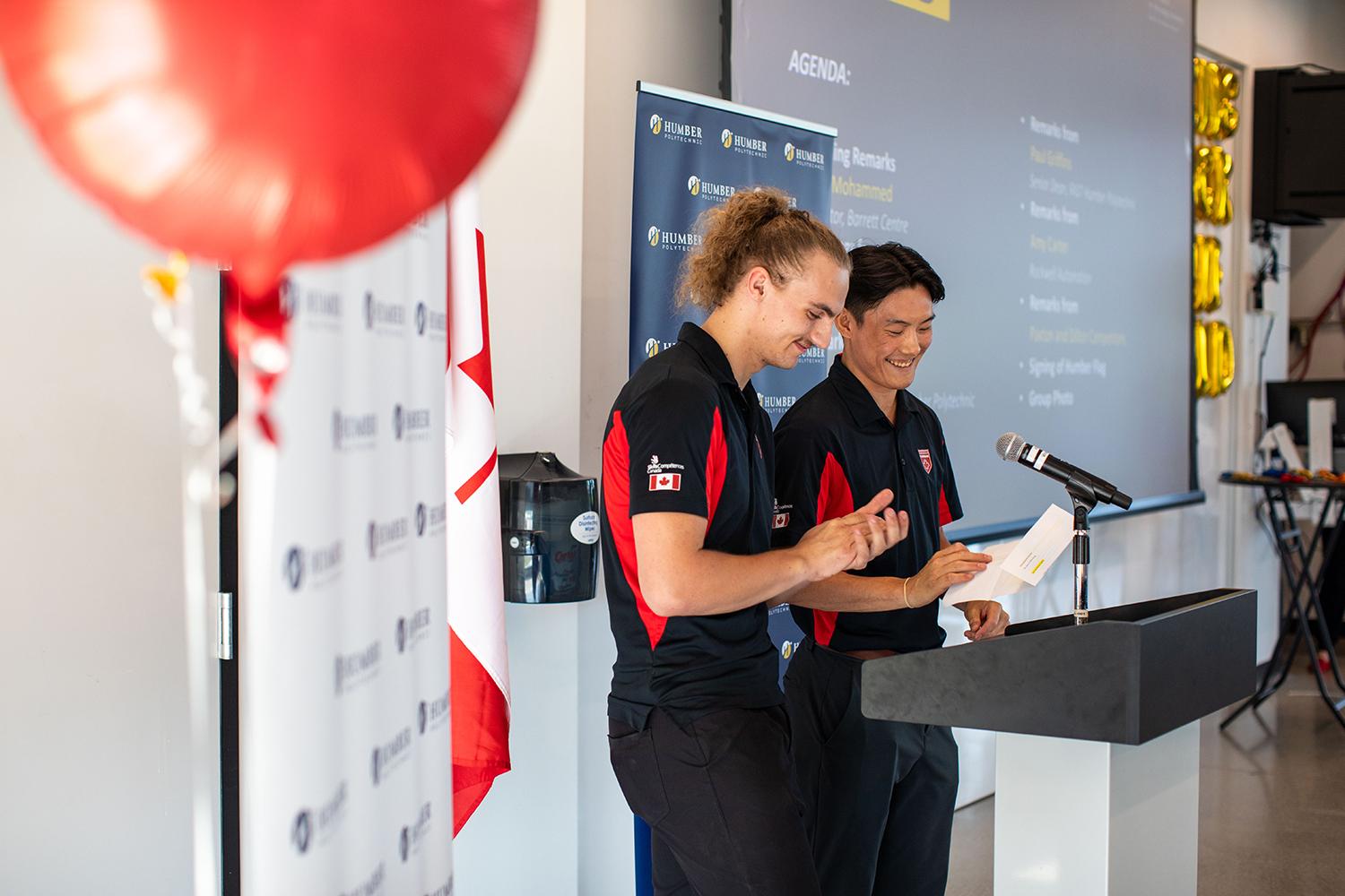 Two smiling people stand at a podium and one is holding a piece of paper in their hand.