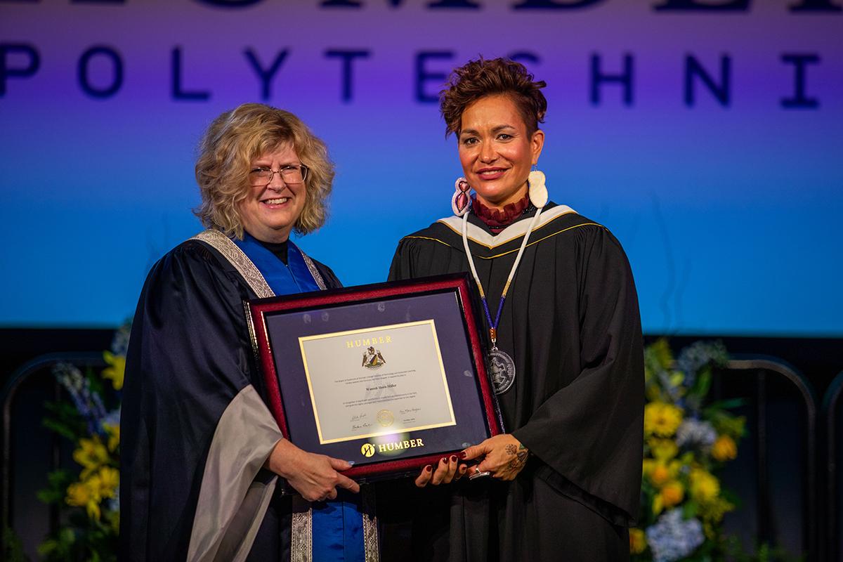 Two smiling people stand together wearing graduation gowns while holding a degree.