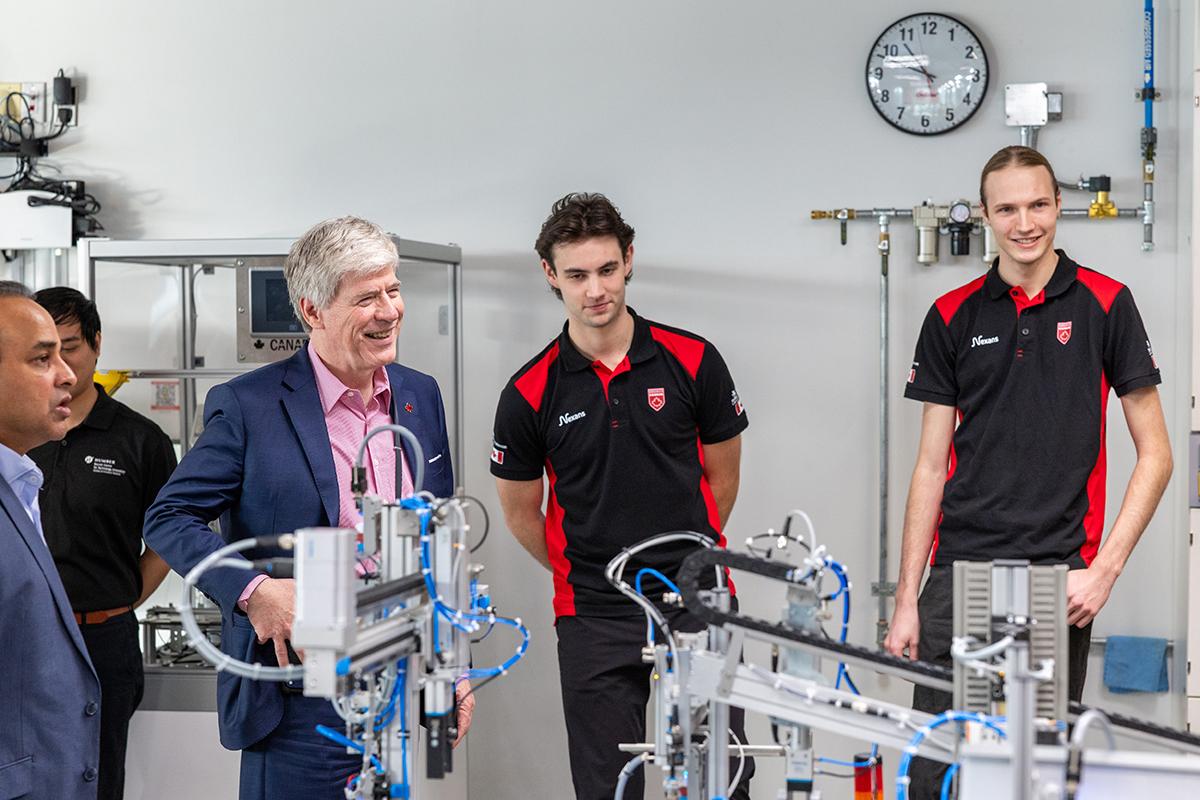 Four people stand together and are talking to each other in front of a mechatronics system.