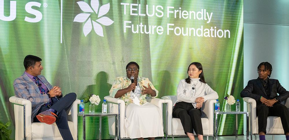 Four people sit in chairs on a stage while one speaks into a microphone. A banner with TELUS Friendly Future Foundation is behin