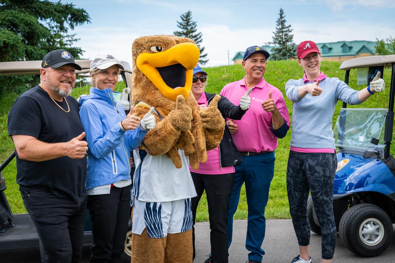 Several people wearing golf attire give a thumb’s up. Standing with them is the Humber Hawks mascot.