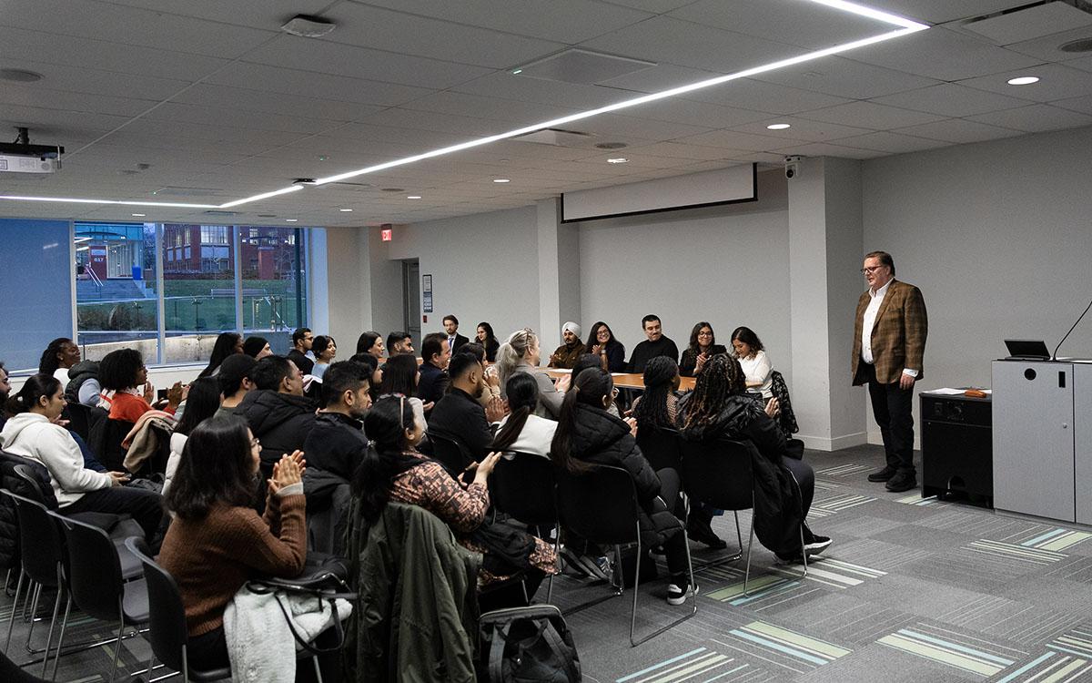 A person standing at the front of a room is speaking to an audience that is watching and listening.