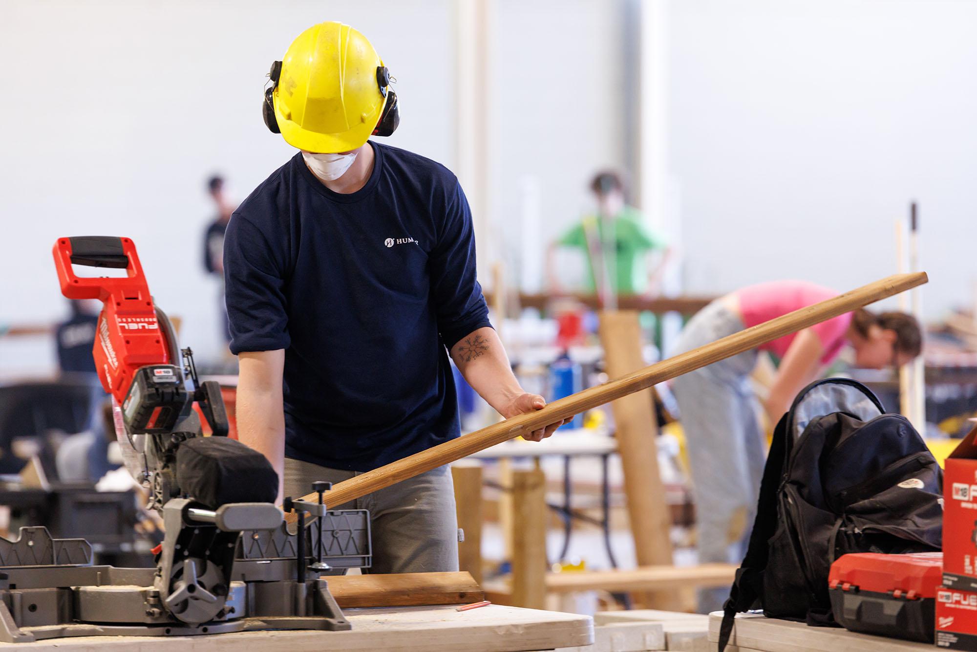 A person wearing a Humber shirt, hard hat, mask and ear protection uses a tool on a piece of wood. 