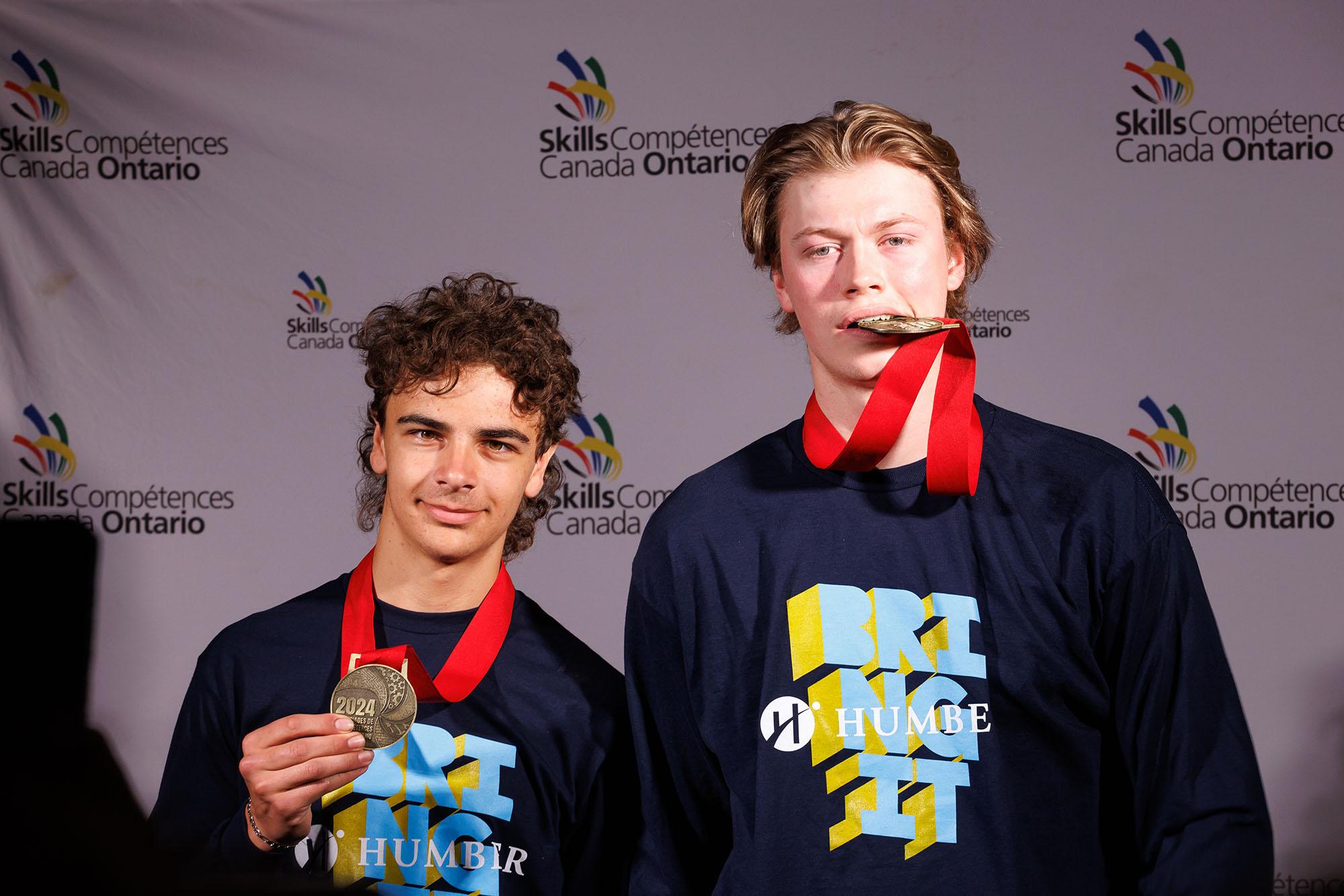 Two people wearing Humber shirts. One is holding up a medal and the other is biting a medal.