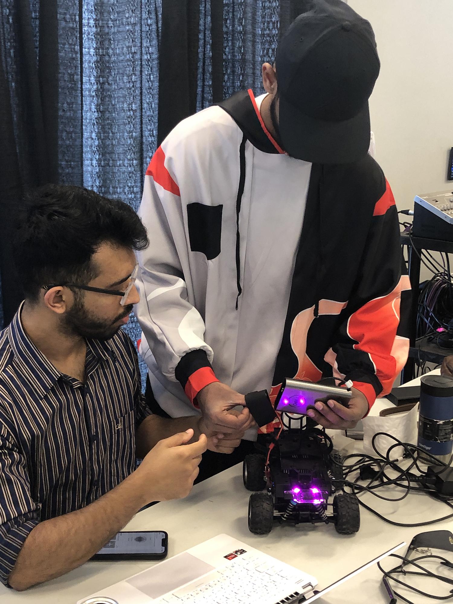 Two people are holding a device while working on a small car that’s sitting on a table.