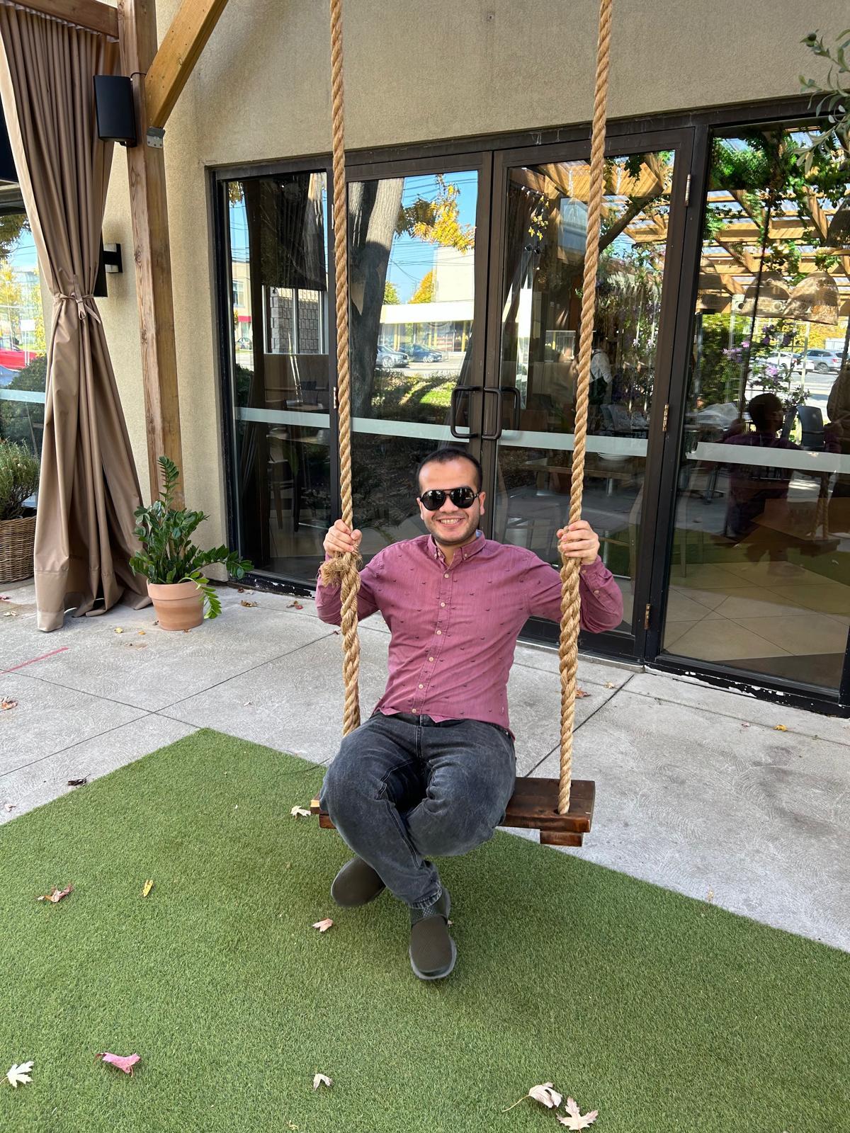 A smiling person is sitting on a swing outside.