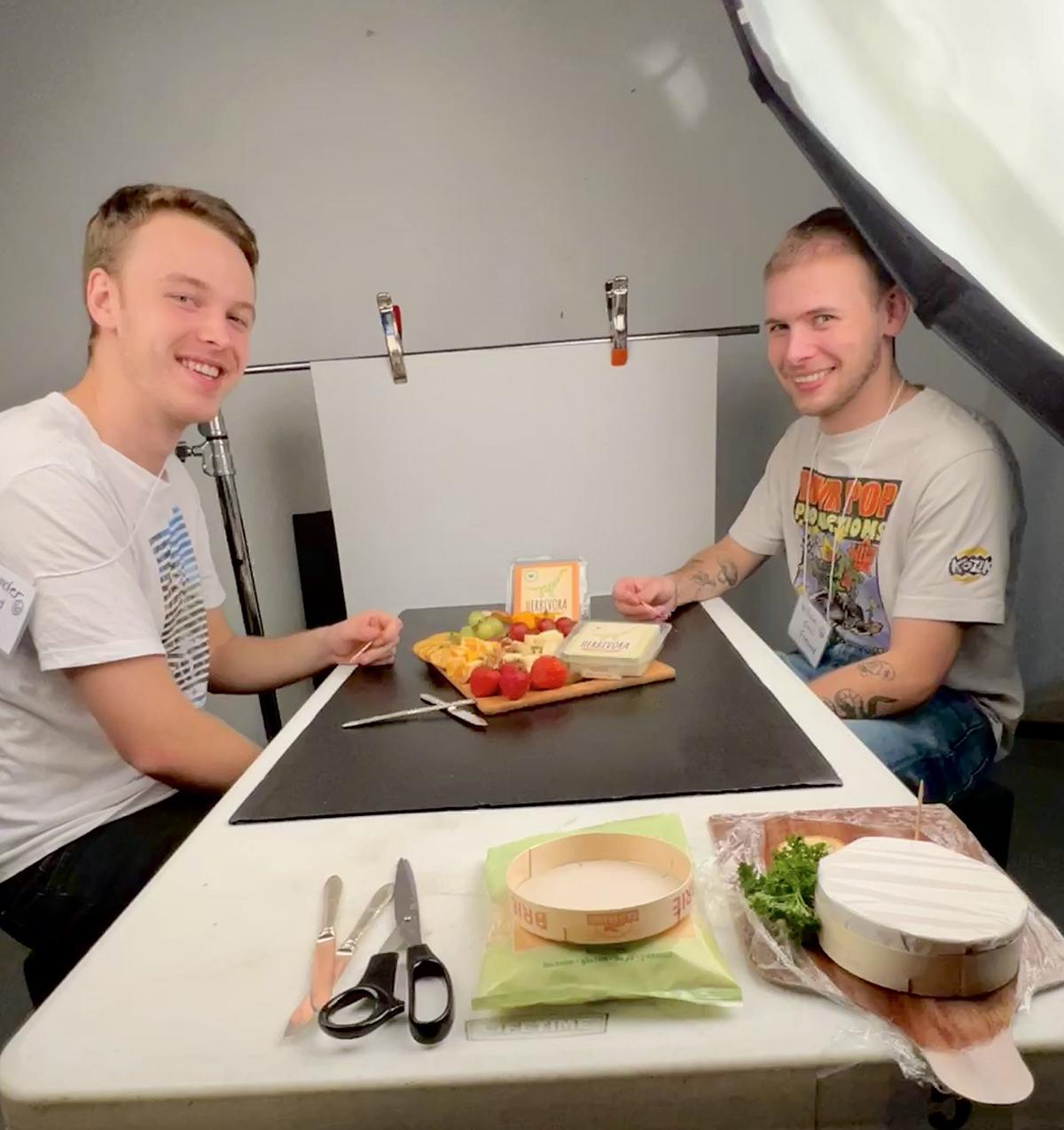 Two people sit at a table that has food arranged on it during a photoshoot.