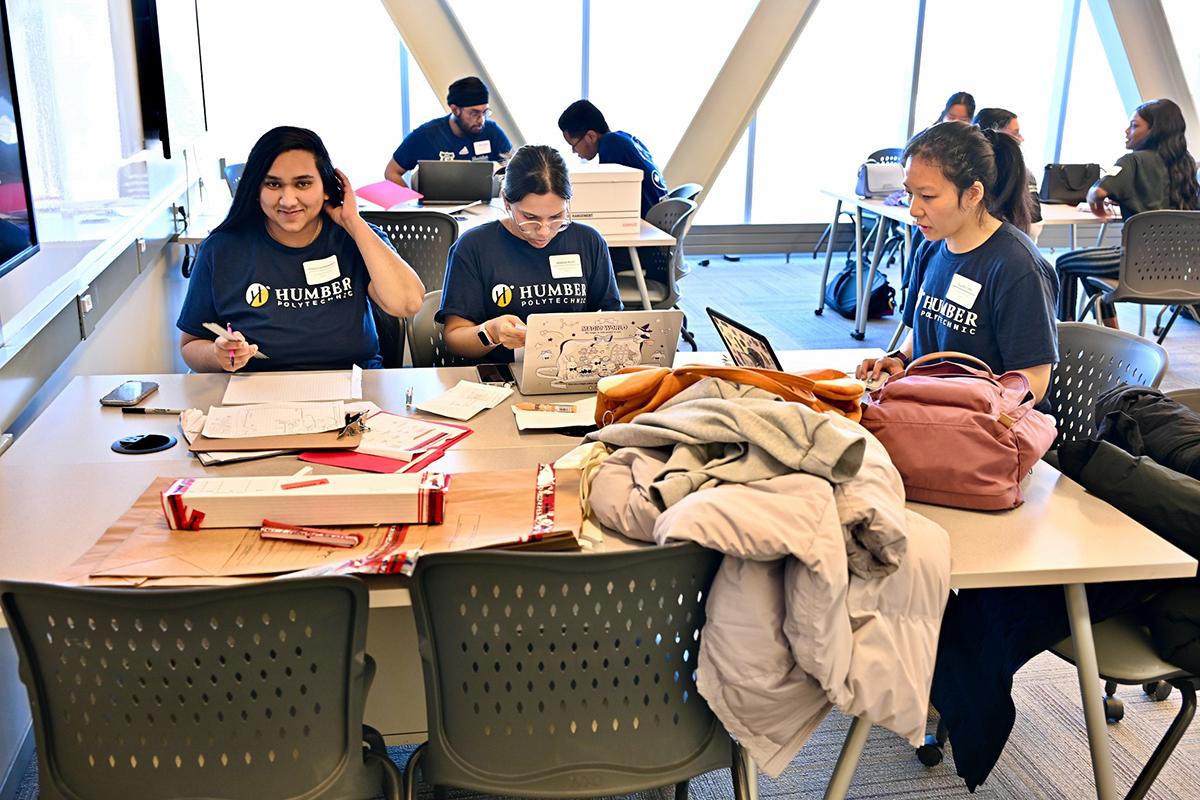 Students wearing Humber Polytechnic shirts are working at a table that has papers and laptops on it.