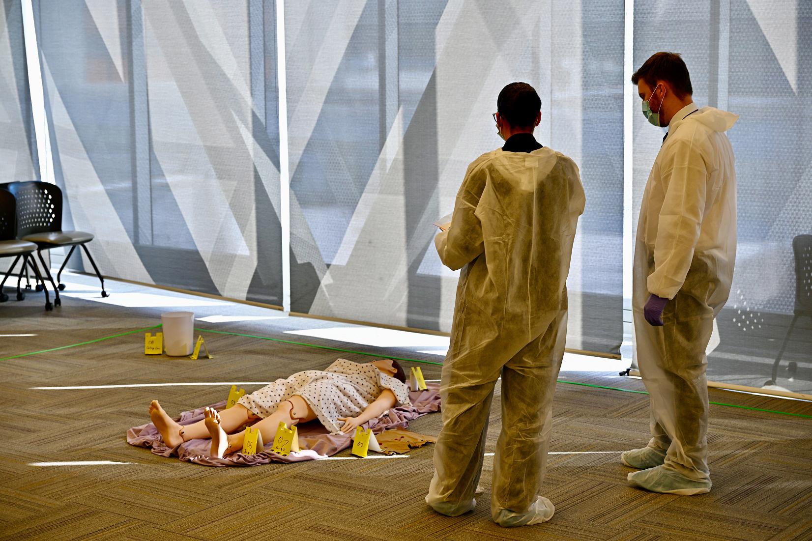 Two people observe a simulated crime scene that has a fake body lying on the ground surrounded by evidence markers.