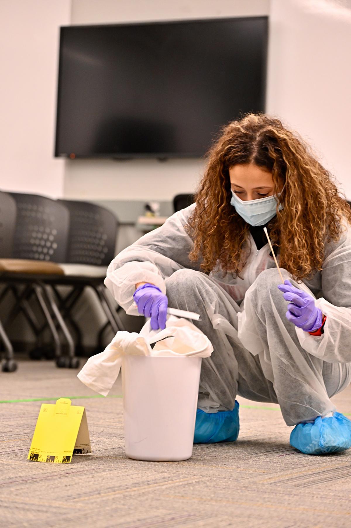 A person wearing a mask, gloves and shoe covers is collecting evidence from a simulated crime scene.