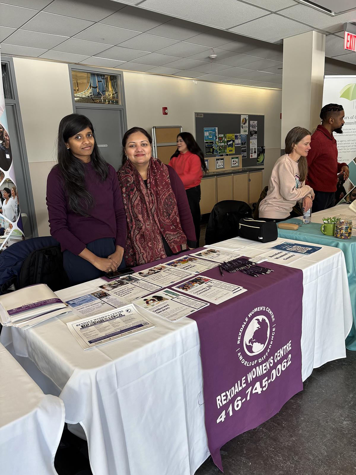 Two people stand at a table with a sign on it that reads Rexdale Women’s Centre.