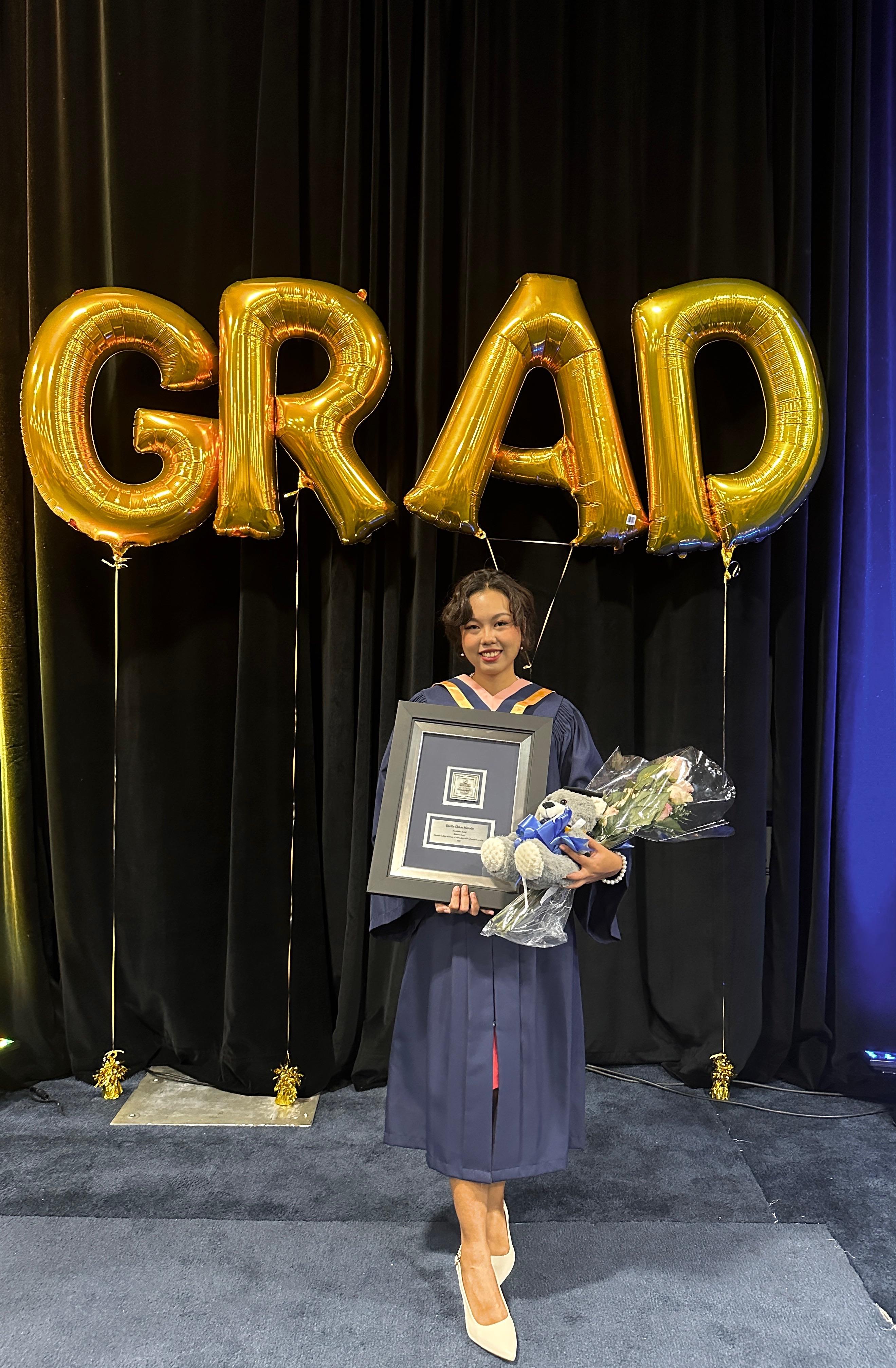 A person wearing a convocation gown holds flowers and a plaque while standing beneath balloons that spell out grad.