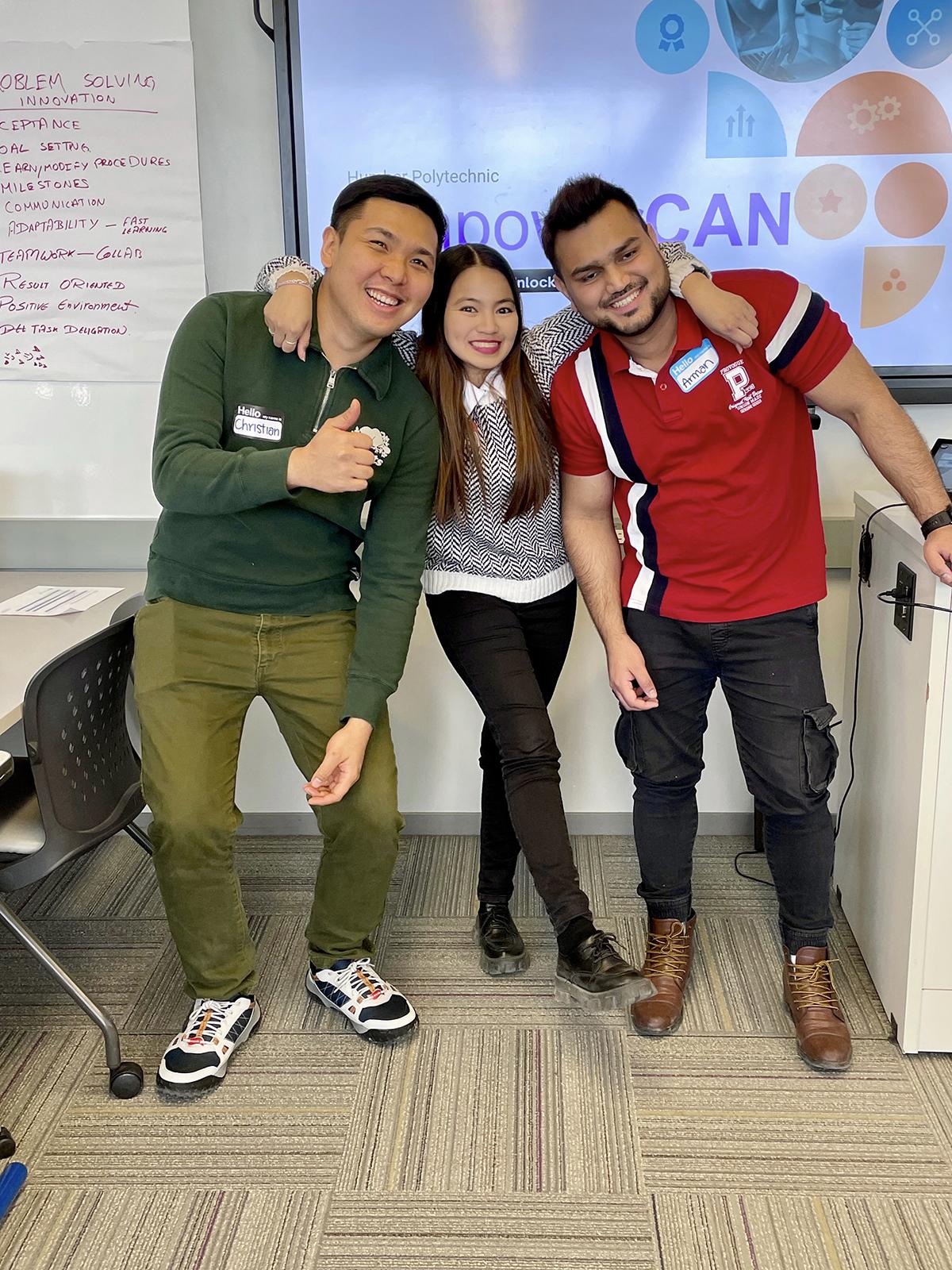 Three smiling people stand together and the person in the middle has their arms over the shoulders of the other two.