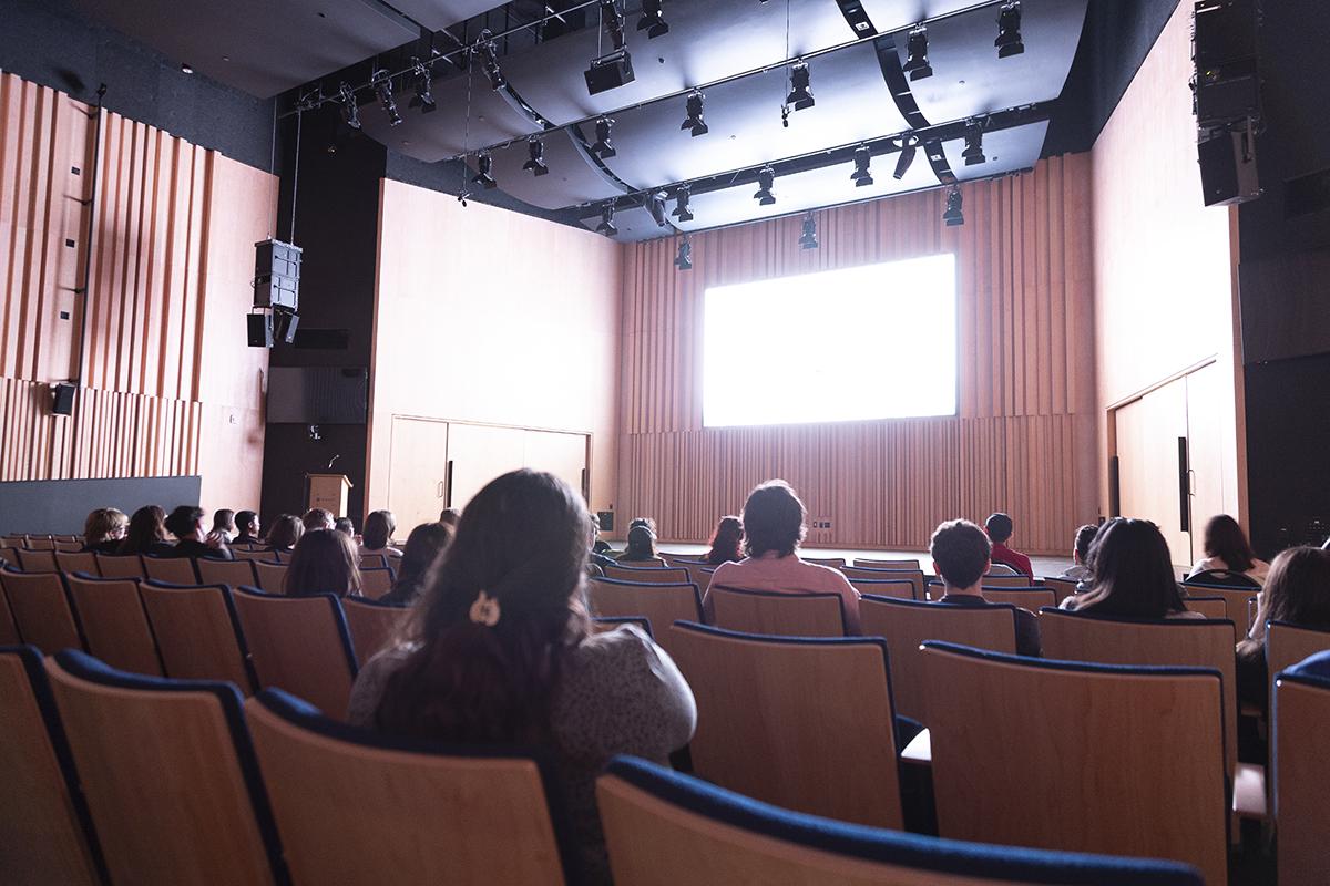 People sitting inside a theatre are watching a screen.