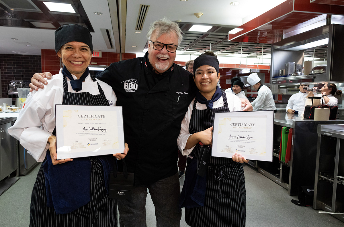 Three smiling people wearing chef’s uniforms stand together in a kitchen. Two of them are holding certificates.