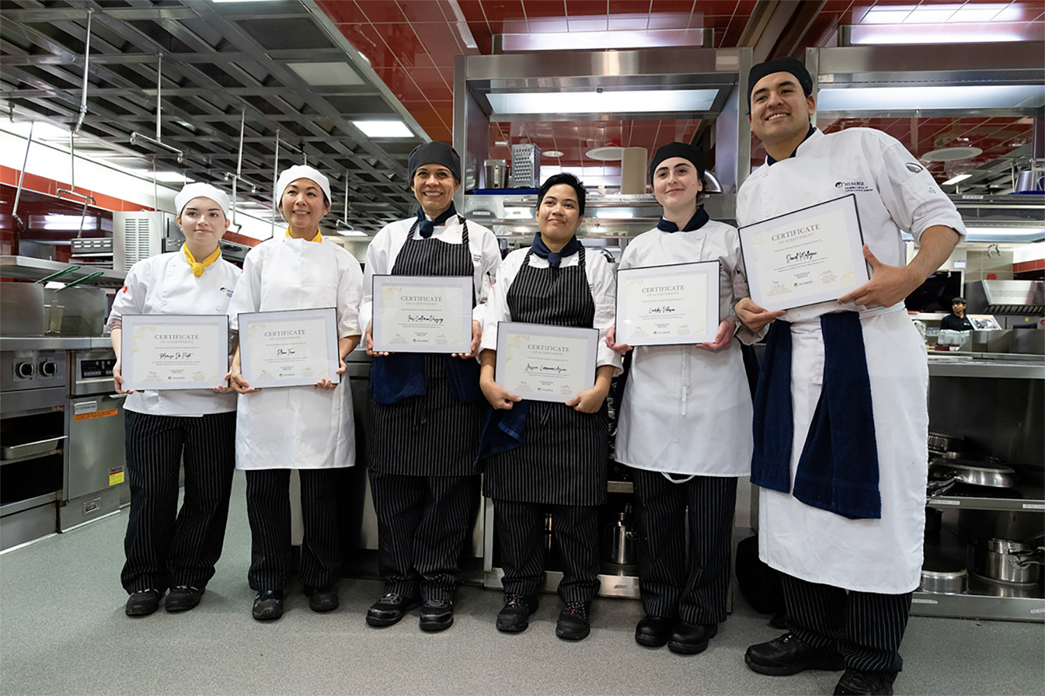 Six people wearing chef’s uniforms are holding certificates as they stand in a kitchen.
