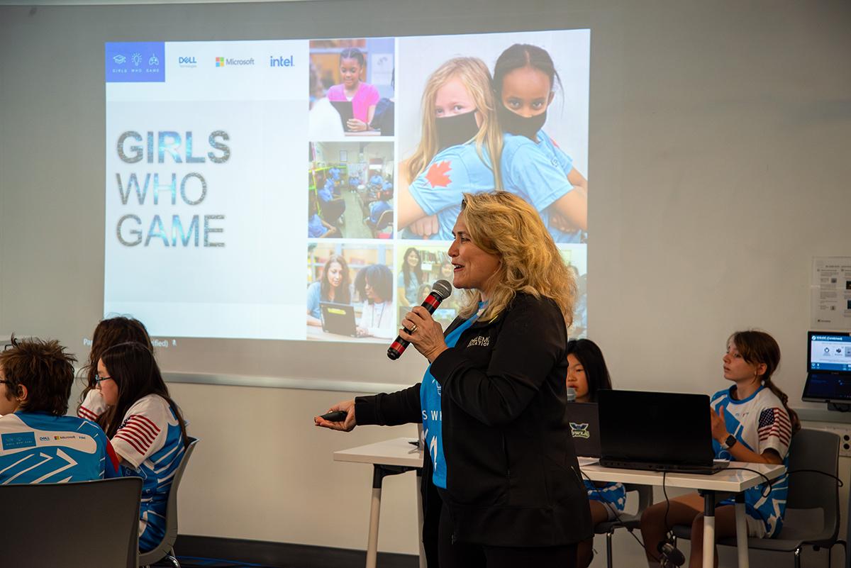 A person speaks into a microphone while standing in front of a screen that has Girls Who Game on it.