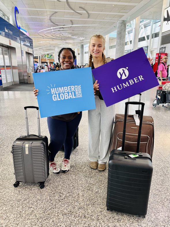 Two people stand next to luggage in an airport holding up signs that read Humber Global Summer School and Humber.