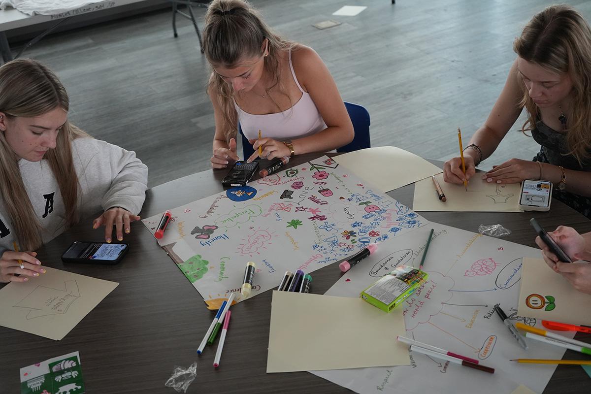 Three people sit at a table that has art supplies on it.