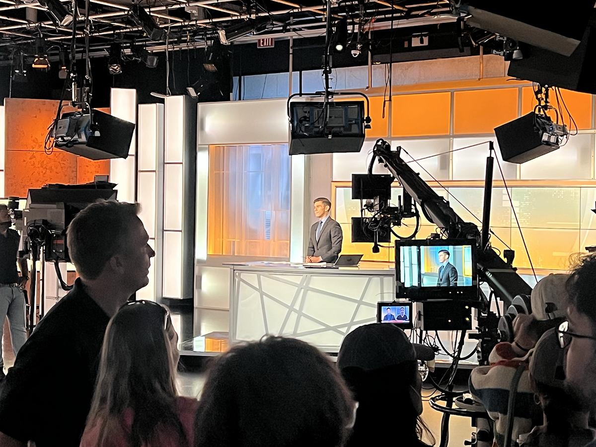 People in a studio watch as a person broadcasts the news.