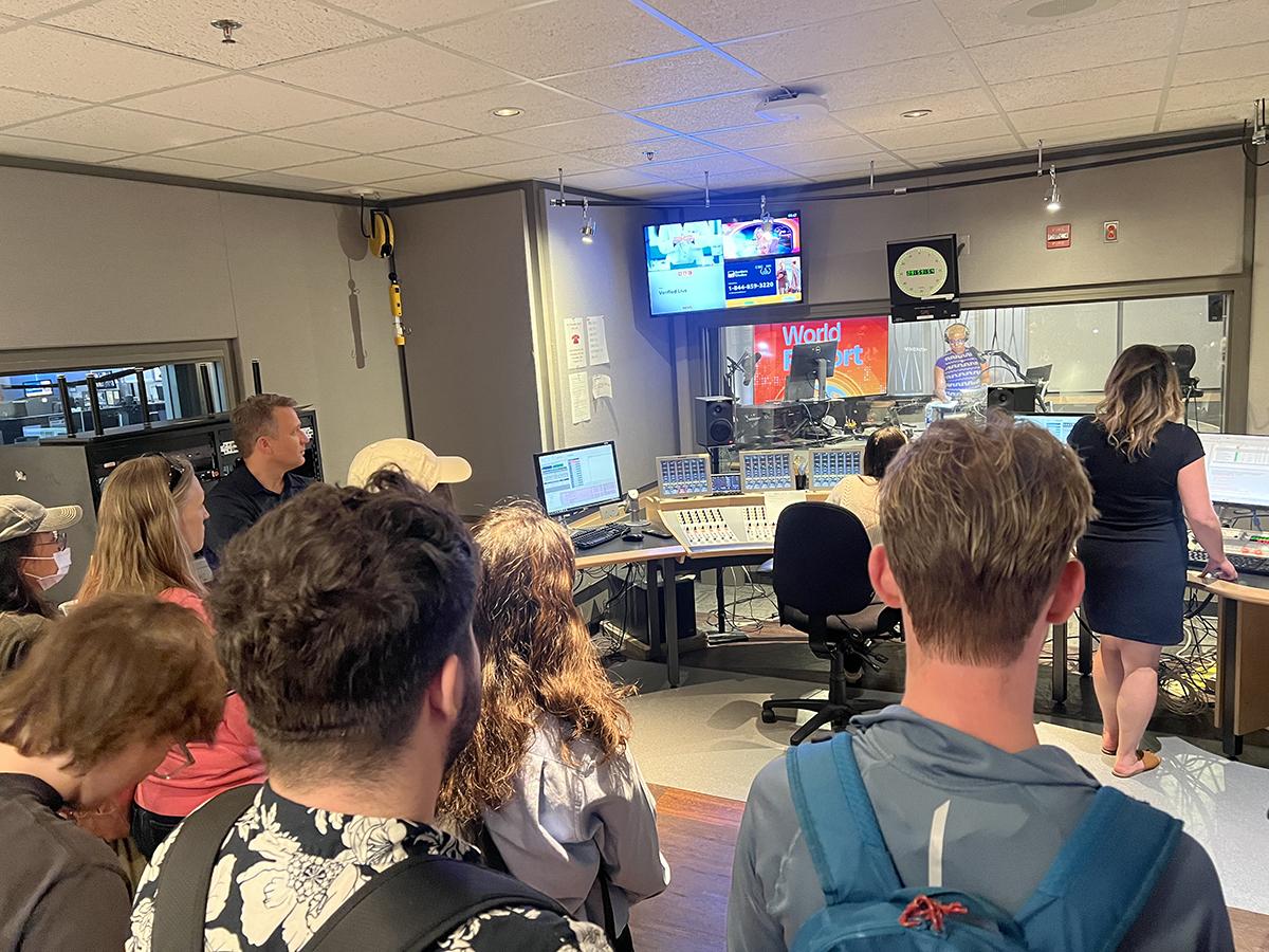 People in a control room watch as a person reads the news in a recording studio.