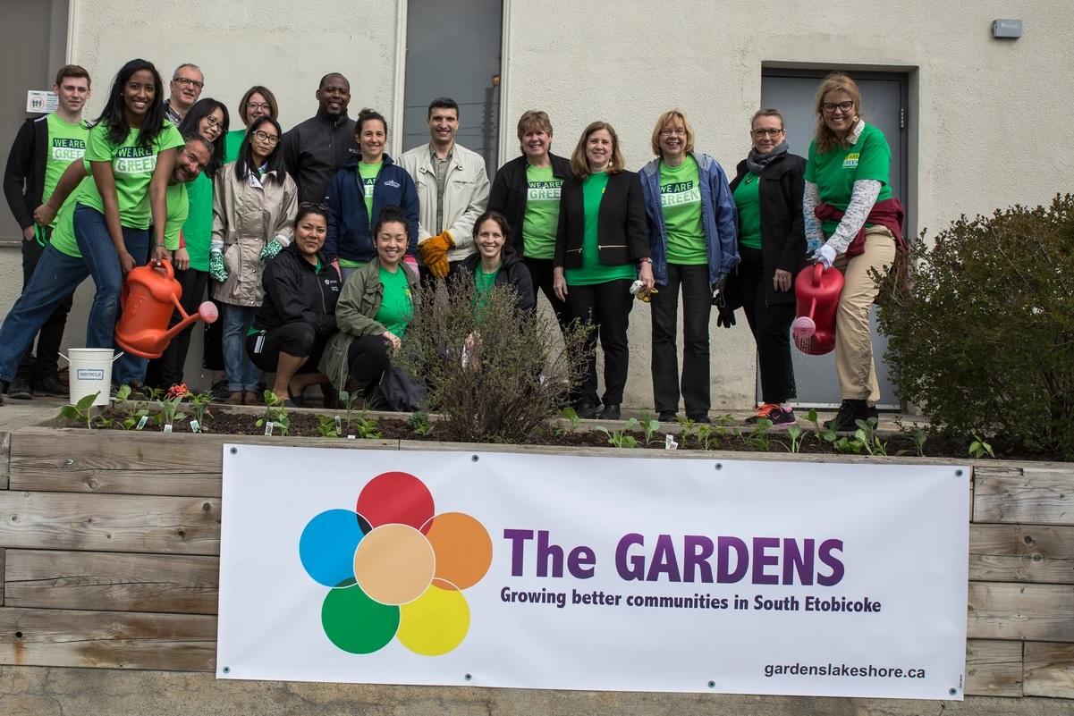 A large group of people stand in front of a banner that reads The Gardens.