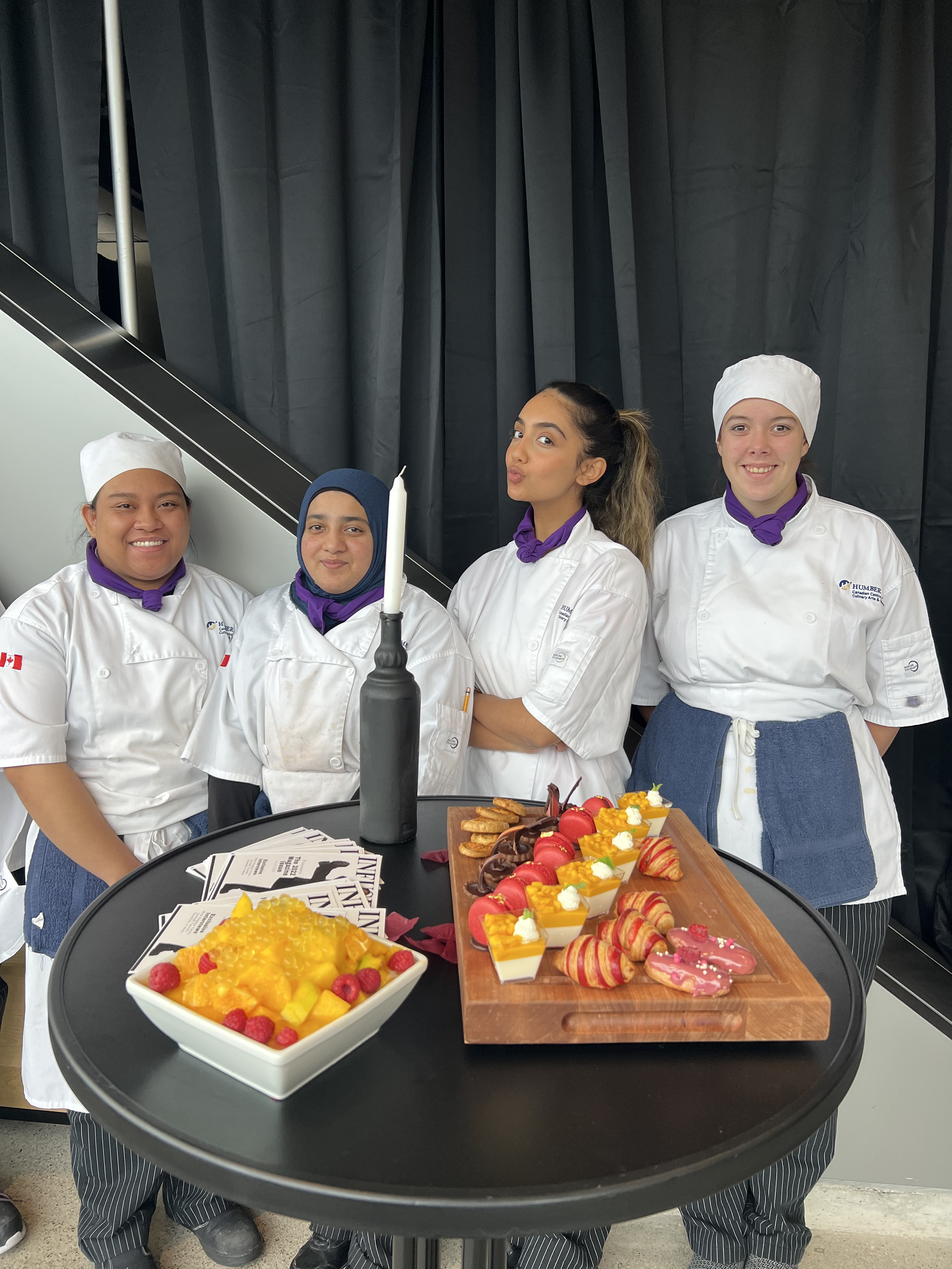 Four Humber College students stand in front of a table that has food on it.